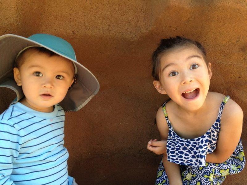 little boy with hat and little girls with patterned dress in front of a brown wall